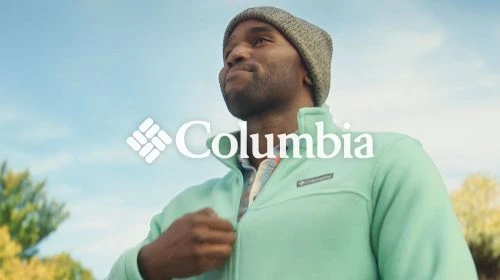 A man in a beanie and fleece jacket stands outside under a blue sky, with the Columbia logo in the foreground. The focus is on the apparel.