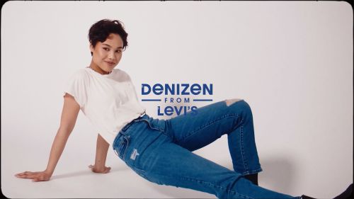 A woman poses indoors on the floor wearing a white t-shirt and blue jeans with a distressed knee, next to the logo for Denizen from Levi's.