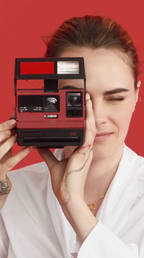 Adult female holds red Polaroid camera in front of face, winking, wearing white lab coat on red seamless background.