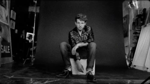 Black and white portrait of a man seated on a box against a dark seamless studio backdrop by Jon Gourlay.