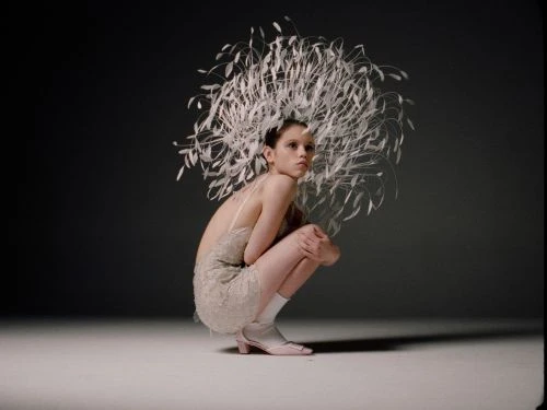 Model squatting in a neutral dress and elaborate white feather headpiece against a dark grey gradient background.