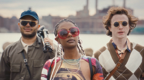 Three people wearing sunglasses pose outdoors near a waterfront. The woman in the middle has braided hair and colorful clothing.