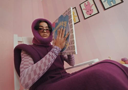 Woman in purple balaclava and jumpsuit reads a book while sitting in a white rocking chair in a pink-toned room with cartoon artwork.