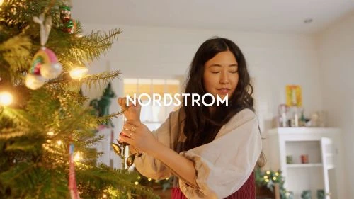 Asian woman decorates Christmas tree with ornaments indoors. Holiday scene features festive lighting, tree decorations, and home decor.