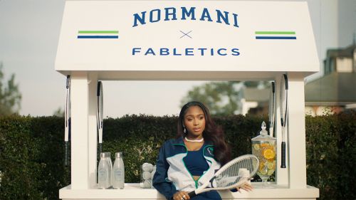 Normani poses with a tennis racket in front of a branded kiosk with water bottles and a citrus drink dispenser in an outdoor setting.
