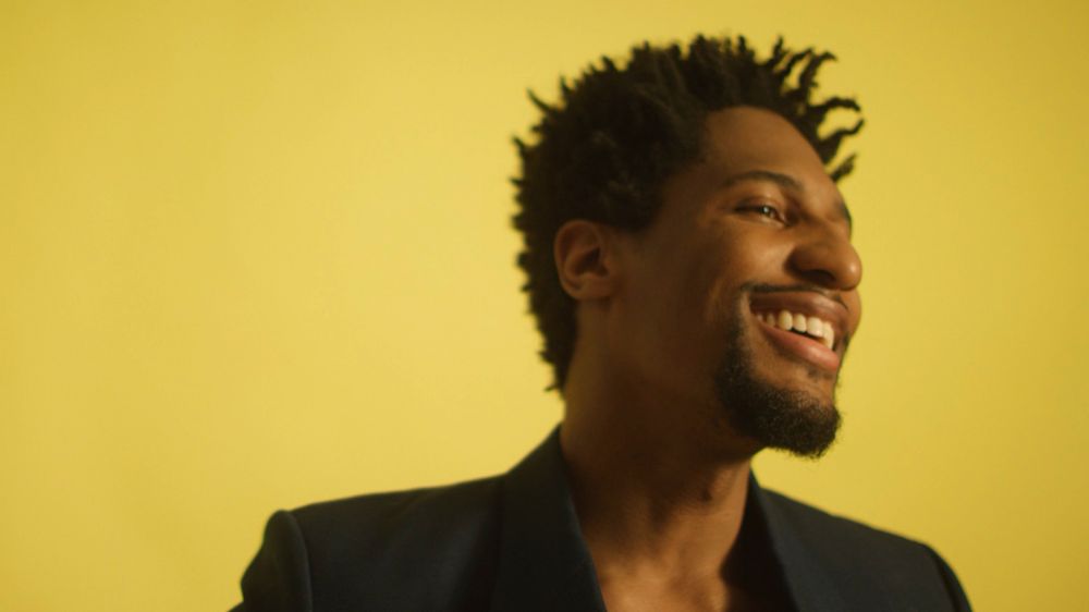 Smiling adult Black man with short hair, wearing a dark blazer, looking to the side against a yellow background in a medium shot.