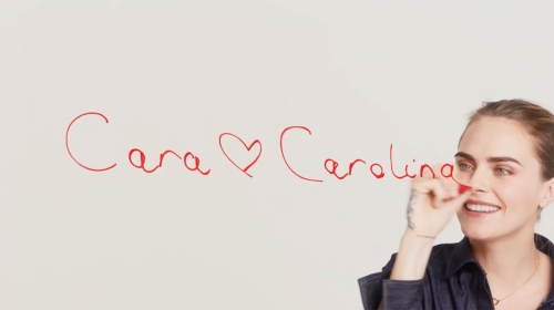Smiling woman in denim shirt with handwritten 'Cara Carolina' on white wall. Candid beauty and handwritten typography.