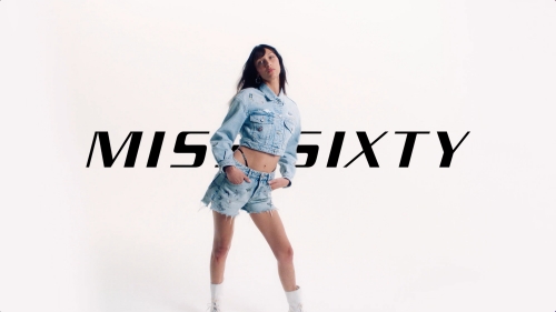 A fashionable young adult woman in denim shorts and jacket strikes a pose in a bright studio setting, showcasing modern casual style.