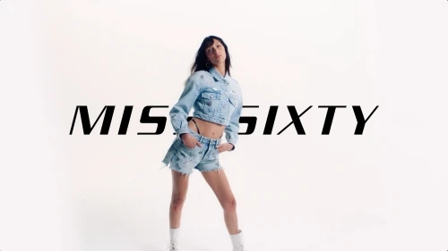 A fashionable young adult woman in denim shorts and jacket strikes a pose in a bright studio setting, showcasing modern casual style.