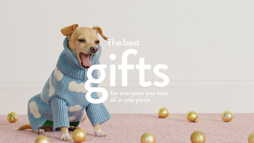 A tan chihuahua wearing a blue sweater with white clouds sits on a pink carpet with gold Christmas ornaments scattered around it in a light-filled room.
