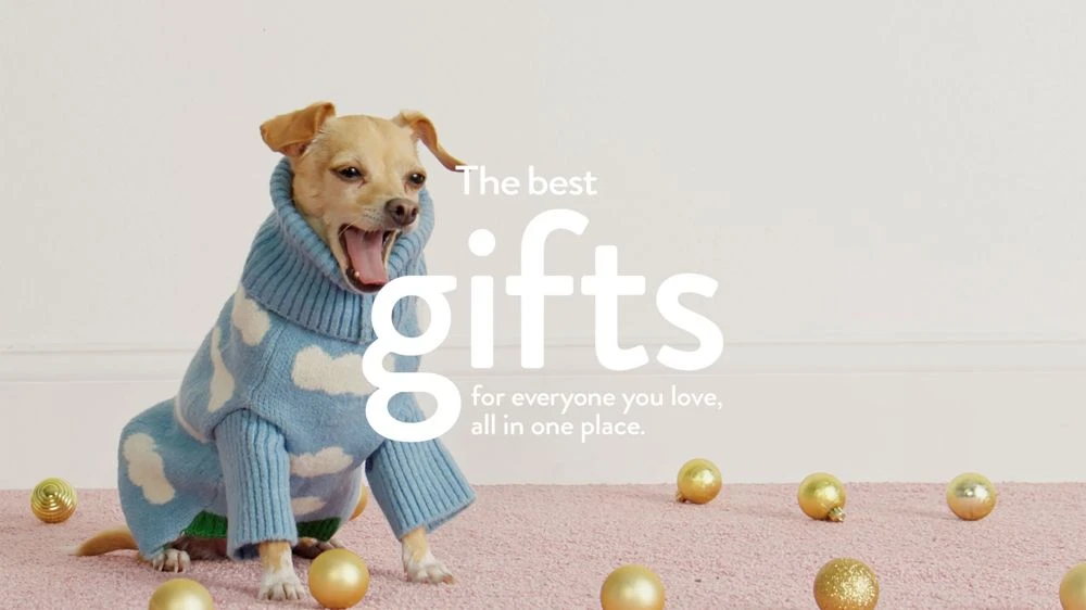 A tan chihuahua wearing a blue sweater with white clouds sits on a pink carpet with gold Christmas ornaments scattered around it in a light-filled room.