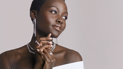Close-up portrait of Lupita Nyong'o wearing diamond drop earrings, layered necklaces, and rings against a neutral studio backdrop.