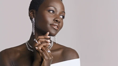 Close-up portrait of Lupita Nyong'o wearing diamond drop earrings, layered necklaces, and rings against a neutral studio backdrop.