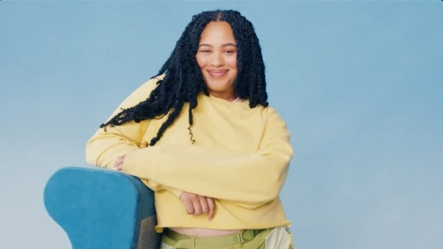 Smiling woman with long dark hair, wearing a yellow cropped sweatshirt and olive pants, leaning on a blue chair against a blue background.
