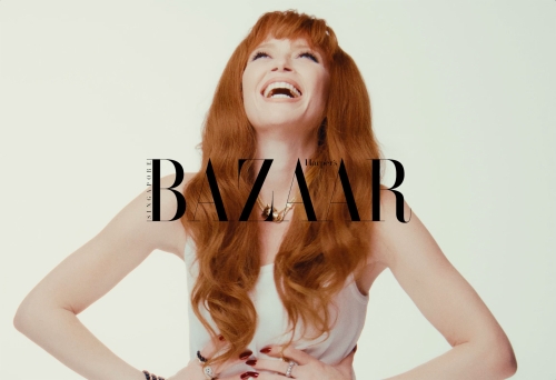 Red-haired woman laughing with head tilted back against a bright white background, wearing a light top and jewellery.