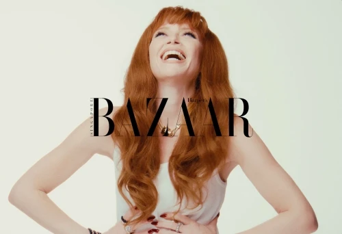 Red-haired woman laughing with head tilted back against a bright white background, wearing a light top and jewellery.