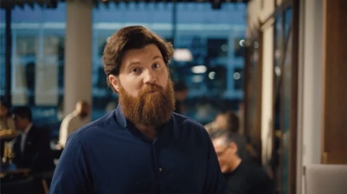 Bearded man in a blue shirt stands indoors at an event with other people and glass windows with natural lighting.