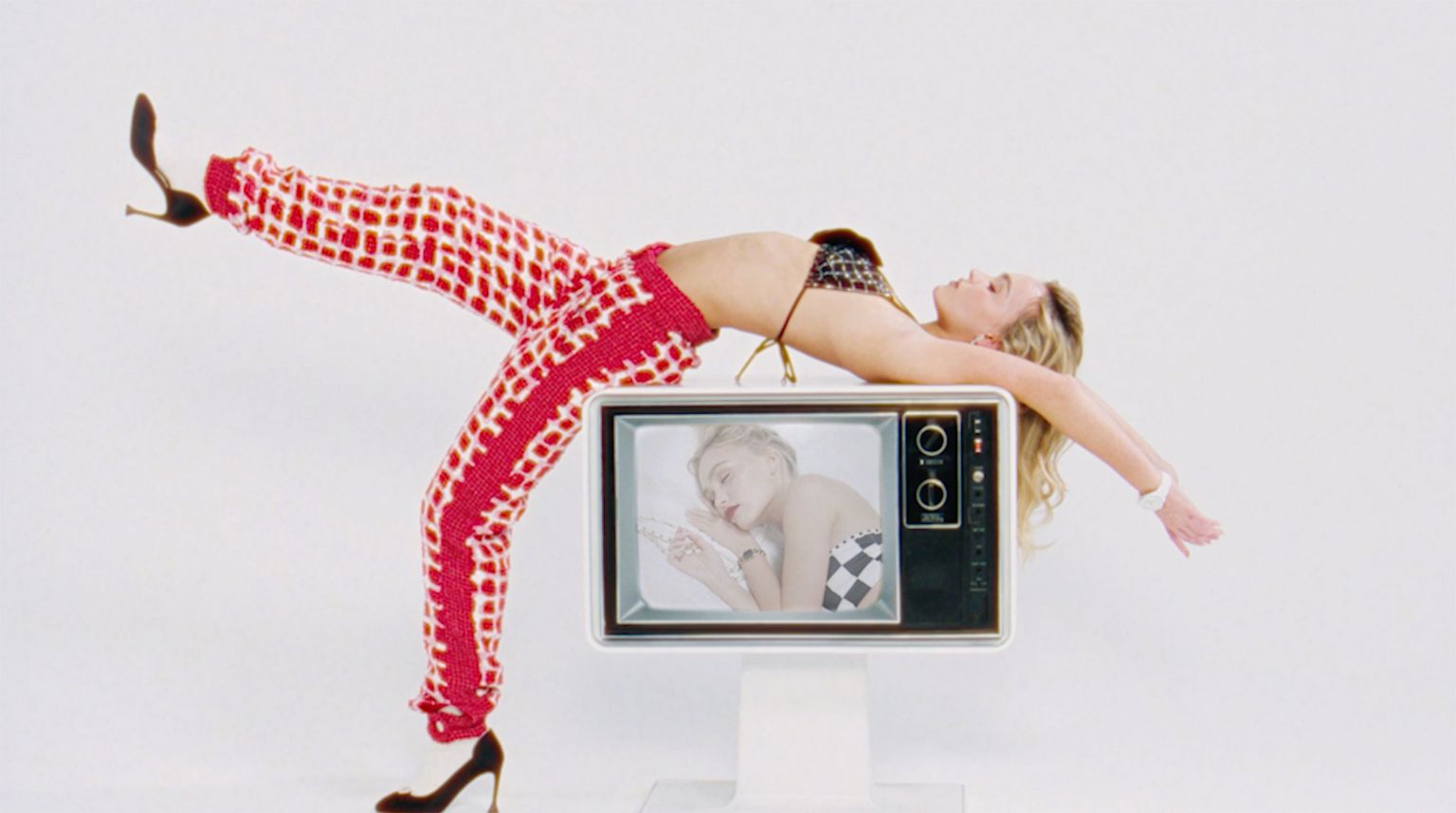 Woman in a bikini top and patterned pants leans back on a retro television in a white photo studio, showcasing a vibrant fashion style.