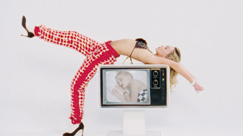 Woman in a bikini top and patterned pants leans back on a retro television in a white photo studio, showcasing a vibrant fashion style.