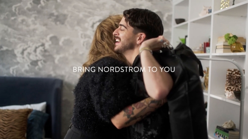 Two people embrace indoors. An adult male with a beard smiles as he hugs an adult female with long hair, with home décor in the background.