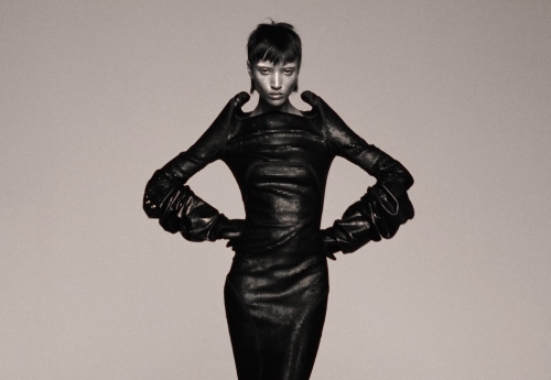 An adult female fashion model in a black dress and gloves poses in a studio. The image has a monochromatic grey color scheme.