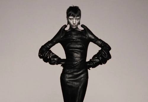 An adult female fashion model in a black dress and gloves poses in a studio. The image has a monochromatic grey color scheme.