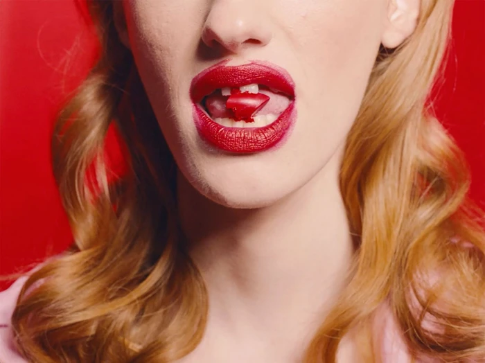 Close-up of woman with blonde hair holding a red lipstick cap in her mouth against a solid red background