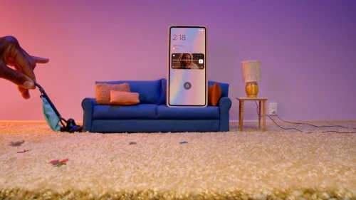 Stylized scene: hand vacuuming carpet near smartphone on sofa. Table lamp, wall, and electrical outlet are also visible.