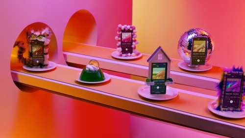 Whimsical still life of smartphones displayed on a yellow platform with colorful props, including a disco ball, a cat figurine, and Jell-O.
