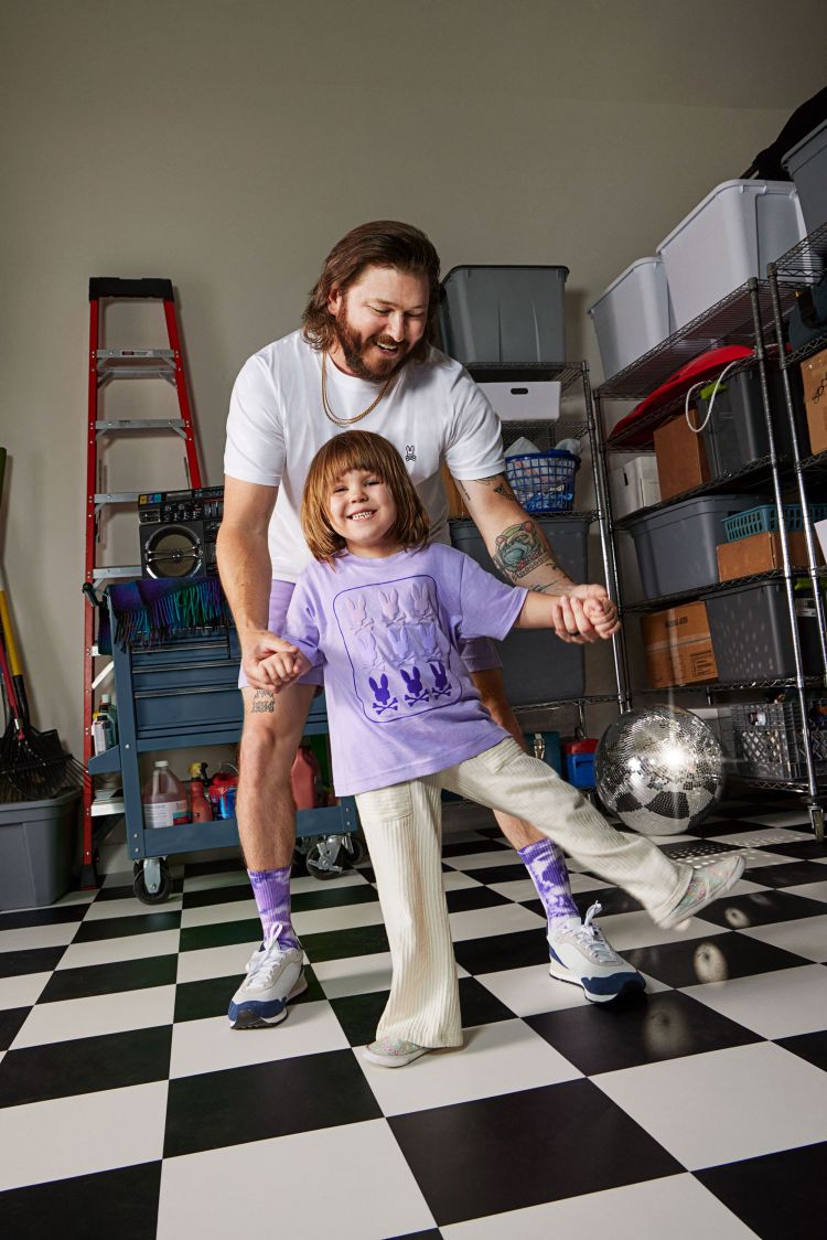 Father and daughter dance in a garage with checkerboard floor. The daughter wears a lavender shirt with white pants.