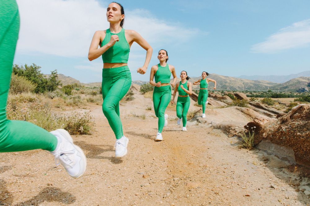 Four adult women running in green athletic wear on a dirt path in a dry, hilly environment with a clear blue sky.