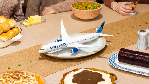 Novel still-life image of an airplane toy placed on a table amidst dinner dishes, vegetables, rolls, and dinnerware on a table runner.