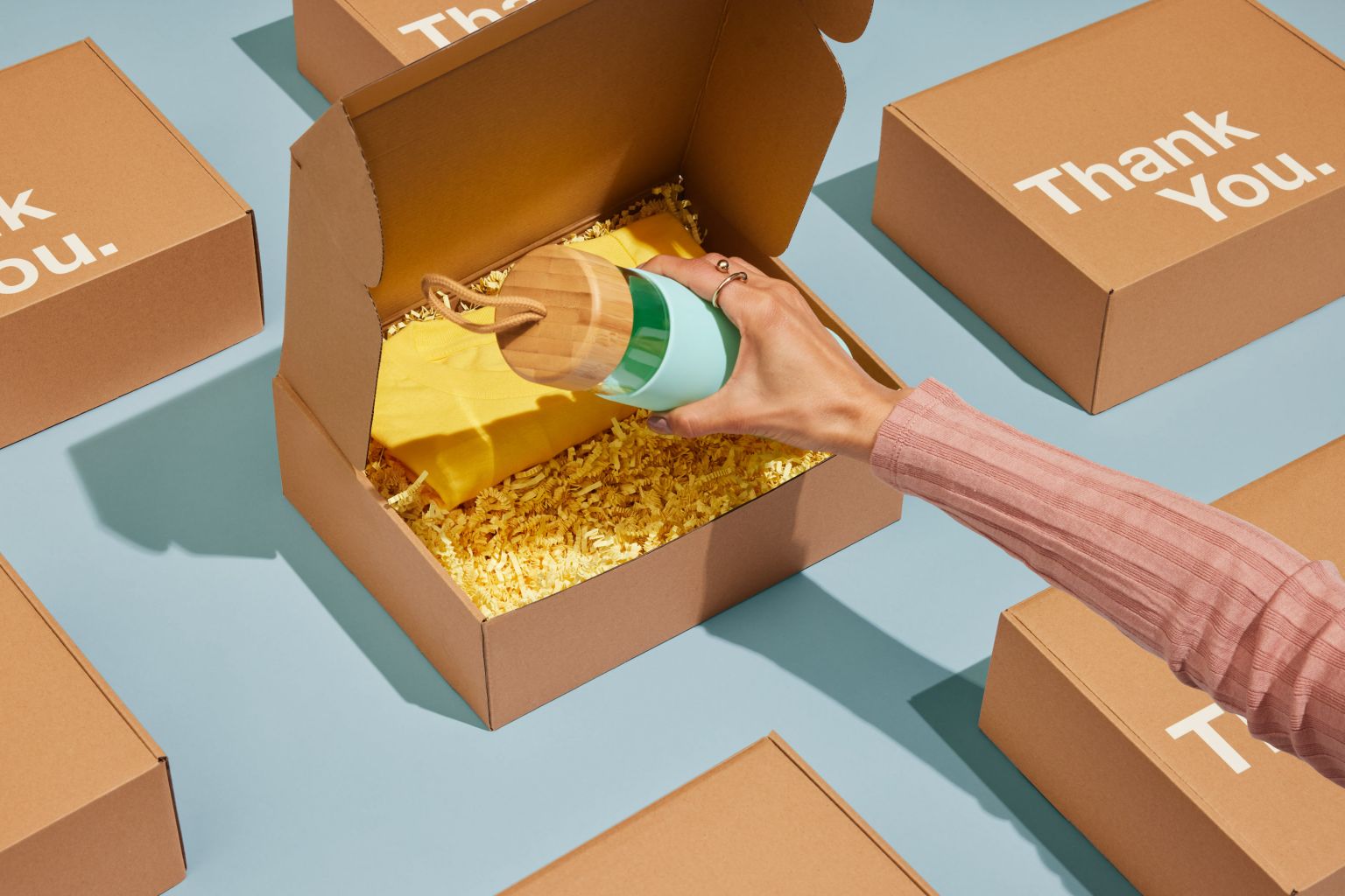Hand placing a light blue and wood grain reusable water bottle into an open tan cardboard box filled with crinkled yellow paper.