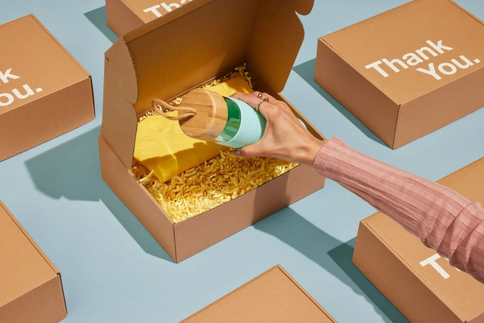 Hand placing a light blue and wood grain reusable water bottle into an open tan cardboard box filled with crinkled yellow paper.