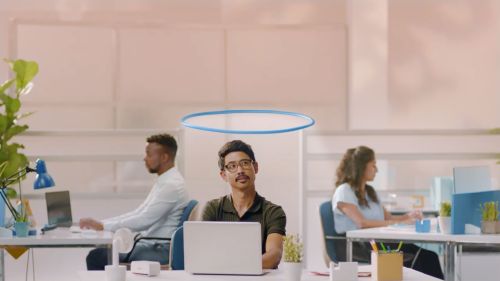 Three office workers sitting at desks with laptops in a bright, modern workspace. An animated halo floats above one person's head.