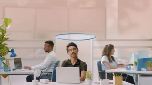 Three office workers sitting at desks with laptops in a bright, modern workspace. An animated halo floats above one person's head.