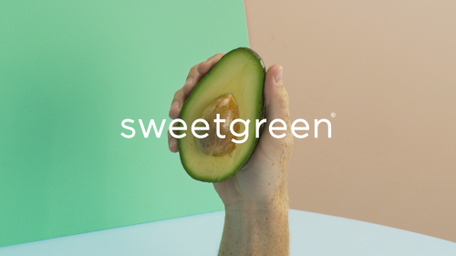 Hand holding a halved avocado in front of a multicolored background with Sweetgreen logo. Food and beverage photography.