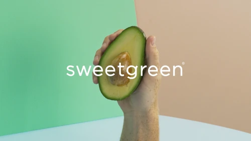 Hand holding a halved avocado in front of a multicolored background with Sweetgreen logo. Food and beverage photography.