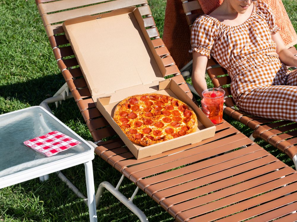 Pepperoni pizza in cardboard box sits on lounge chair with woman in gingham, refreshing drink, and grass in a sunlit outdoor scene.