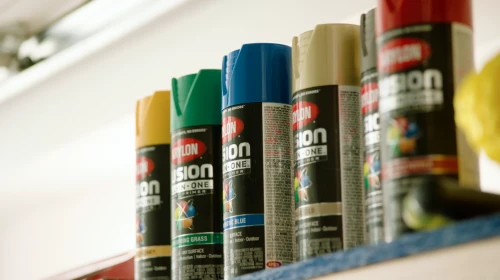 Row of colorful Fusion All-In-One spray paint cans on a shelf, showcasing shades of yellow, green, blue, and red in a tidy display.