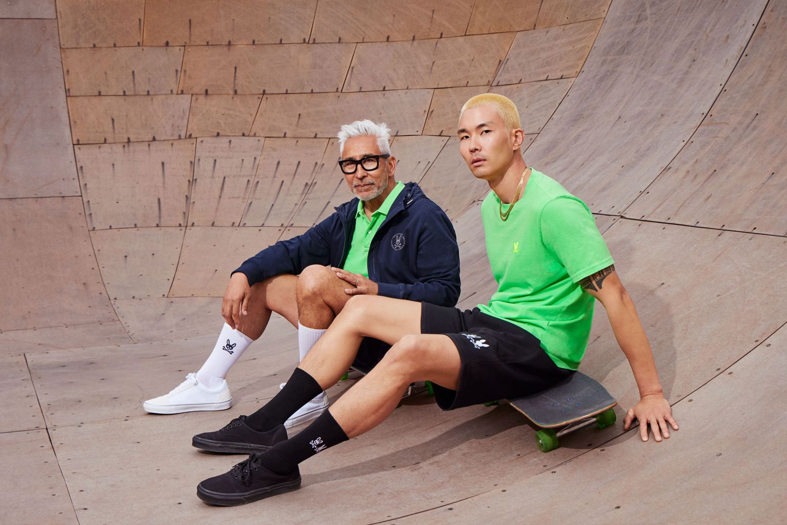 Two stylish men in a skatepark wearing streetwear apparel, one sitting on a skateboard, showcasing a blend of casual and sporty fashion.