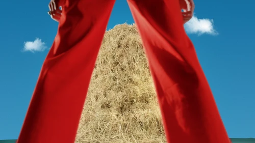 Haystack with blue sky and cloud background is seen between the legs of a person wearing bright red pants.