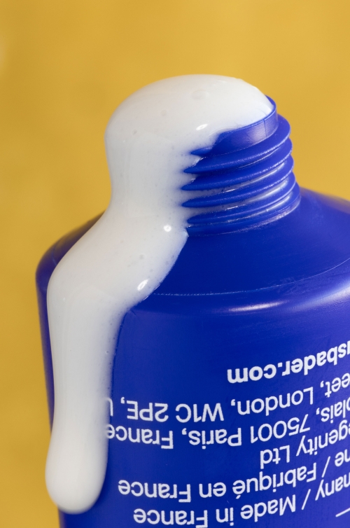 Close-up of a blue and white skincare tube with a swirl of white cream coming out of the top against a solid yellow background.