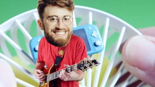 Smiling man in red t-shirt plays electric guitar; video game console and white fan are also visible in a green background studio.