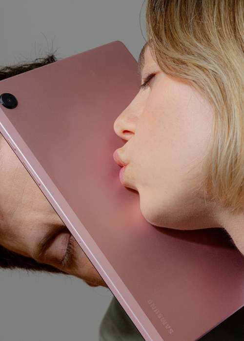 Conceptual shot of a woman and man facing each other and kissing a pink tablet screen in a studio setting with neutral background.