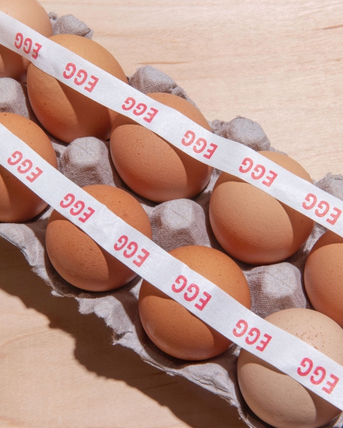 Brown eggs in a grey carton are stacked, bisected by a white tape with red 'EGG' text, all sitting on a light wood surface.