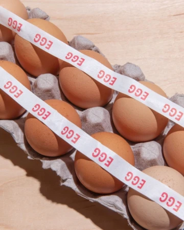 Brown eggs in a grey carton are stacked, bisected by a white tape with red 'EGG' text, all sitting on a light wood surface.