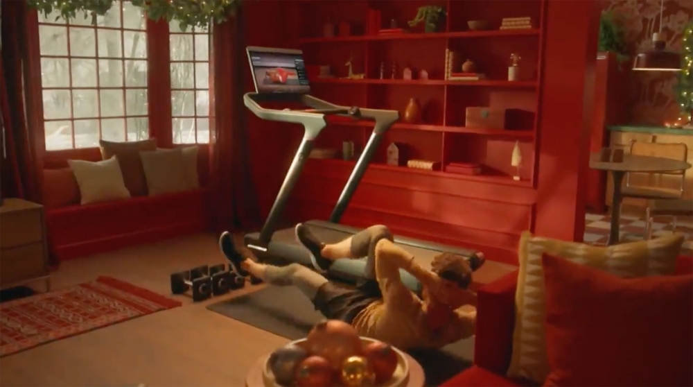 A man is seen doing sit-ups on a mat near a treadmill in a warmly decorated room, with red and gold accents, showcasing a fitness routine.