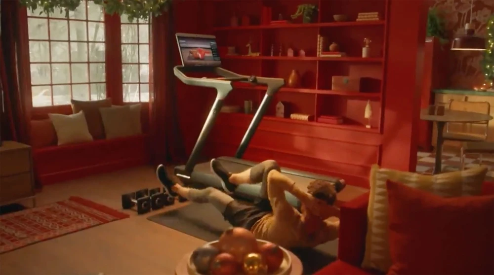 A man is seen doing sit-ups on a mat near a treadmill in a warmly decorated room, with red and gold accents, showcasing a fitness routine.
