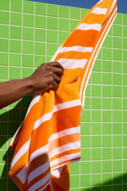 Close-up of a dark-skinned arm holding an orange and white striped towel against a bright green tiled wall in sunlight.